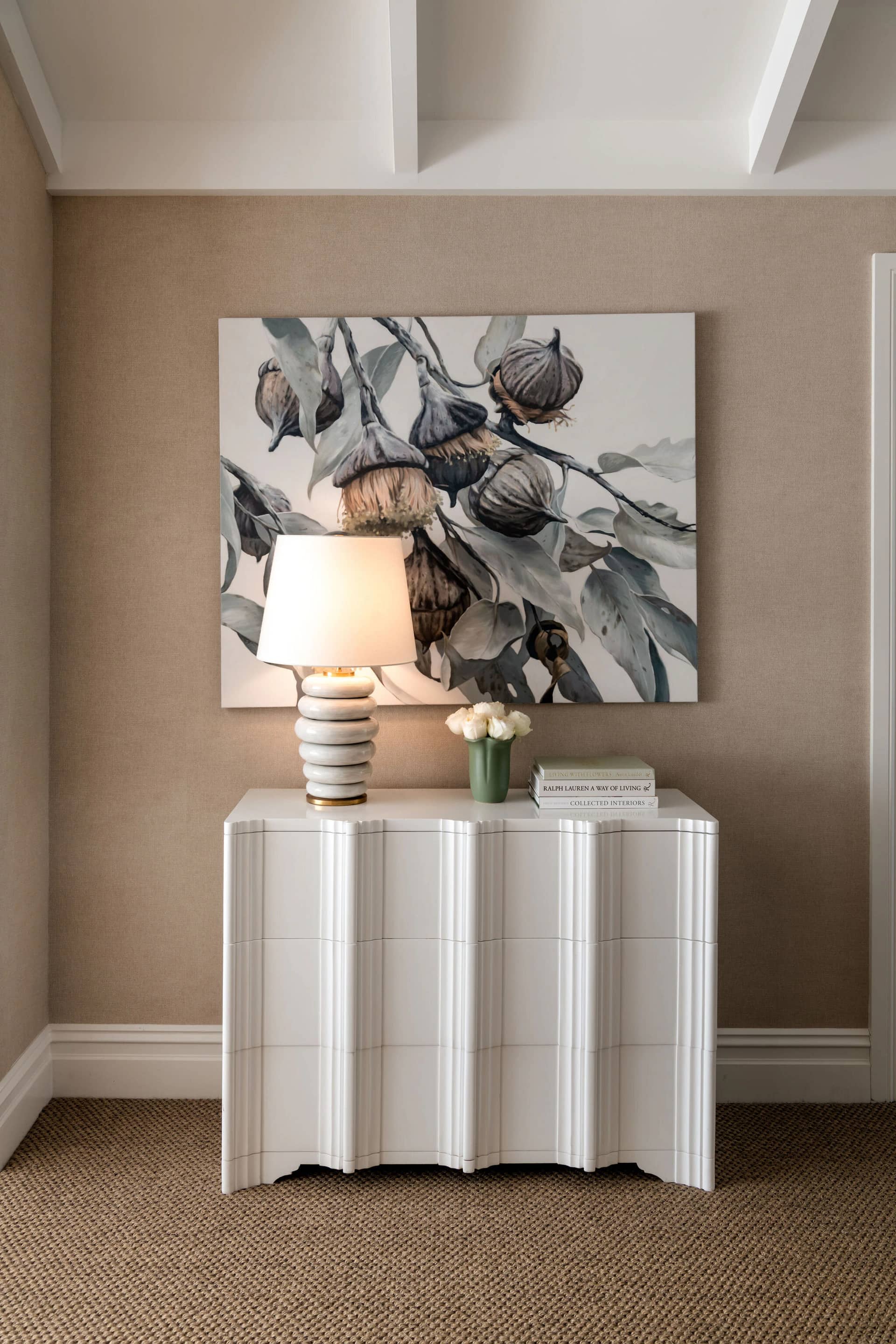 A bedroom corner with a scalloped side table, lamp and botanical artwork
