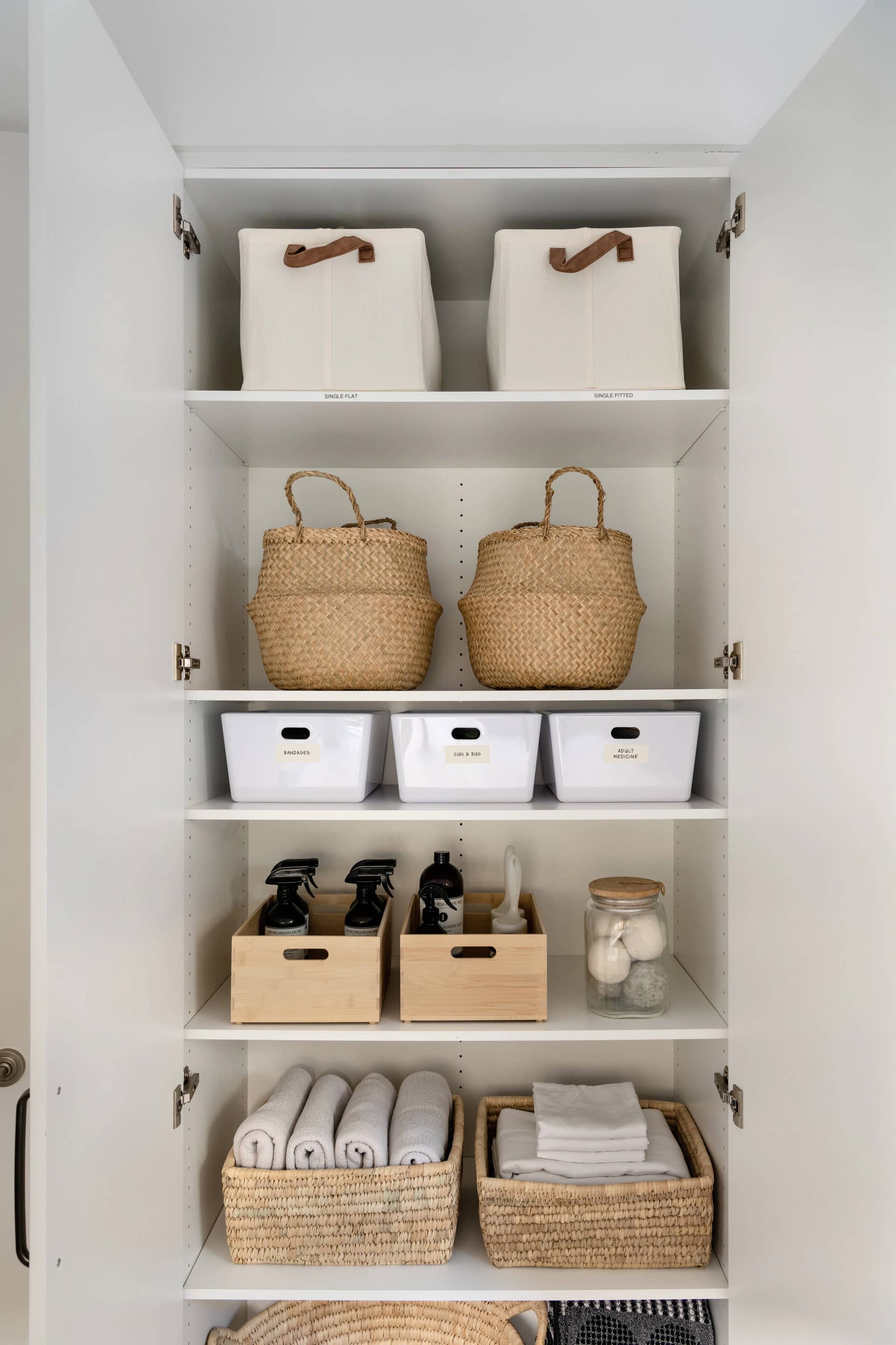 A linen cupboard with white bins, woven baskets and folded towels