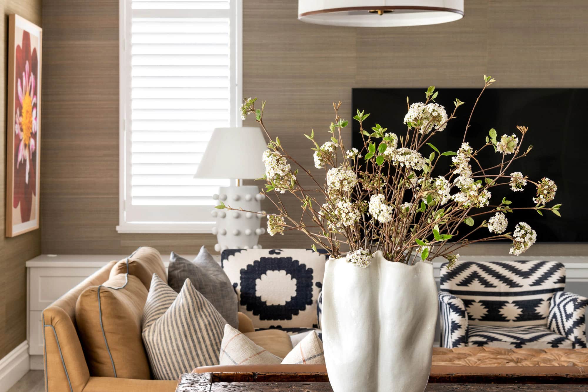 A styled living room with a white sculptural vase of blossom on a rustic timber console, a tan sofa and a drum pendant light